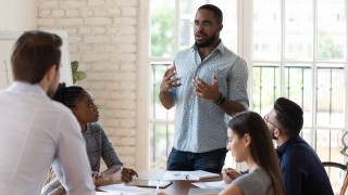 Serious black businessman manager boss mentor talk to diverse staff people teaching interns at corporate briefing table explaining project strategy at multiracial company office meeting in boardroom.