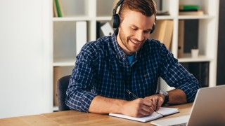 portrait of smiling man in headphones making notes while taking part in webinar at tabletop with laptop in office