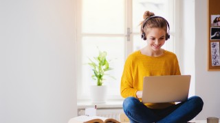 A young happy college female student sitting at the table at home, using headphones and laptop when studying.